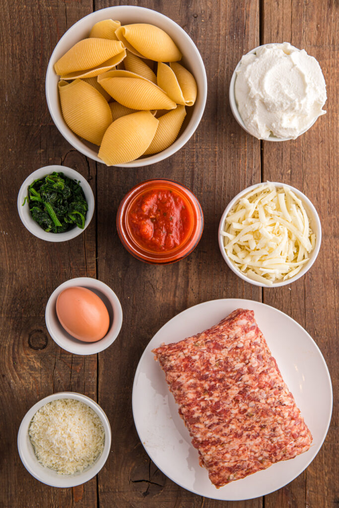 top down image showing uncooked jumbo pasta shells, ricotta cheese, spinach, red sauce, mozzarella, parmesan, an egg, and sausage