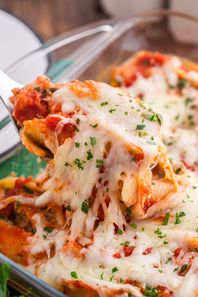 Top down image showing a glass baking dish filled with pasta shells that are topped with red sauce and cheese. One shell is being pulled out of the pan with a white serving spoon