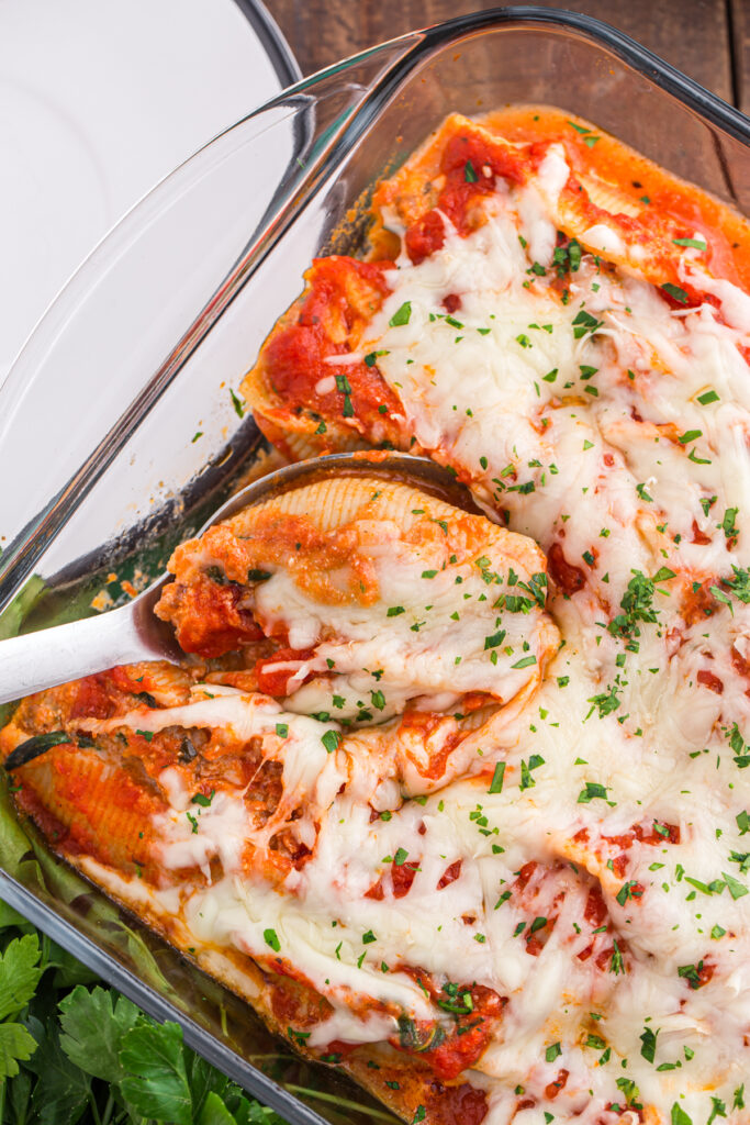Top down image showing a glass baking dish filled with pasta shells that are topped with red sauce and cheese. One shell is being pulled out of the pan with a white serving spoon