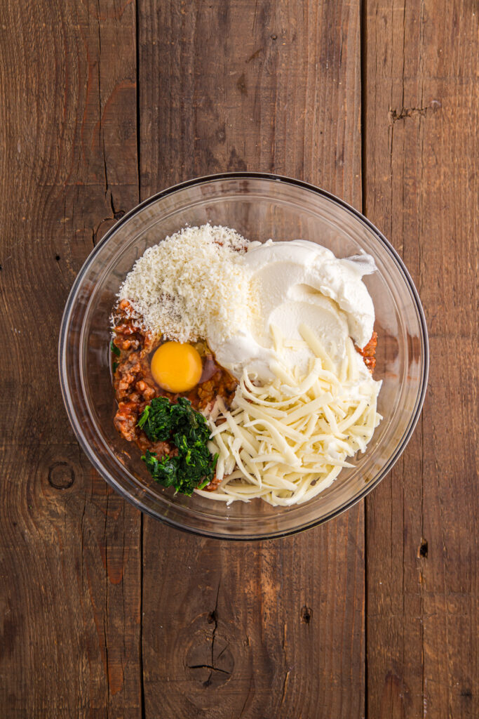 top down image showing a glass mixing bowl with ricotta, mozzarella, parmesan, egg, and spinach