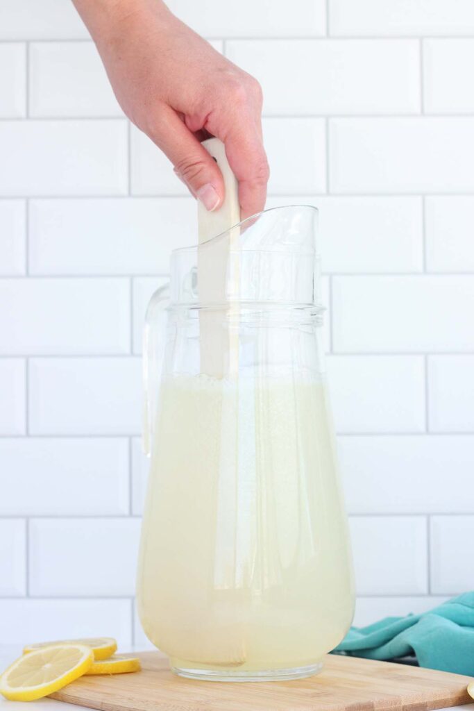 Lemonade being stirred with a wooden spoon inside a glass pitcher