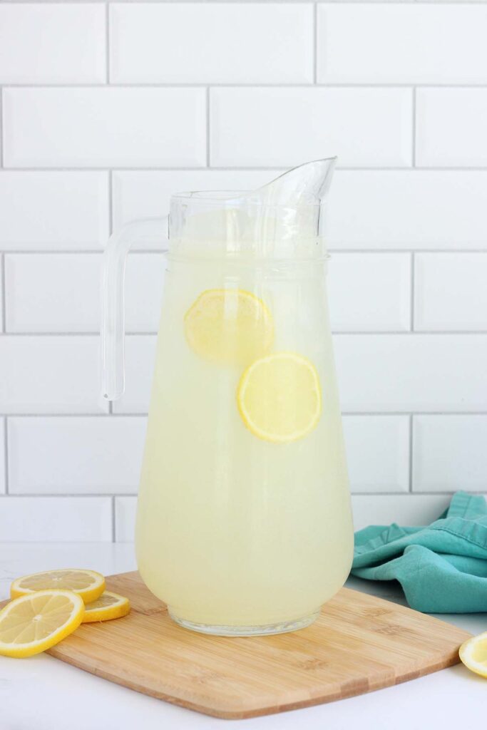 pitcher filled with lemonade, ice and two lemon slices sitting on a cutting board with more lemon slices and a teal napkin off to the side
