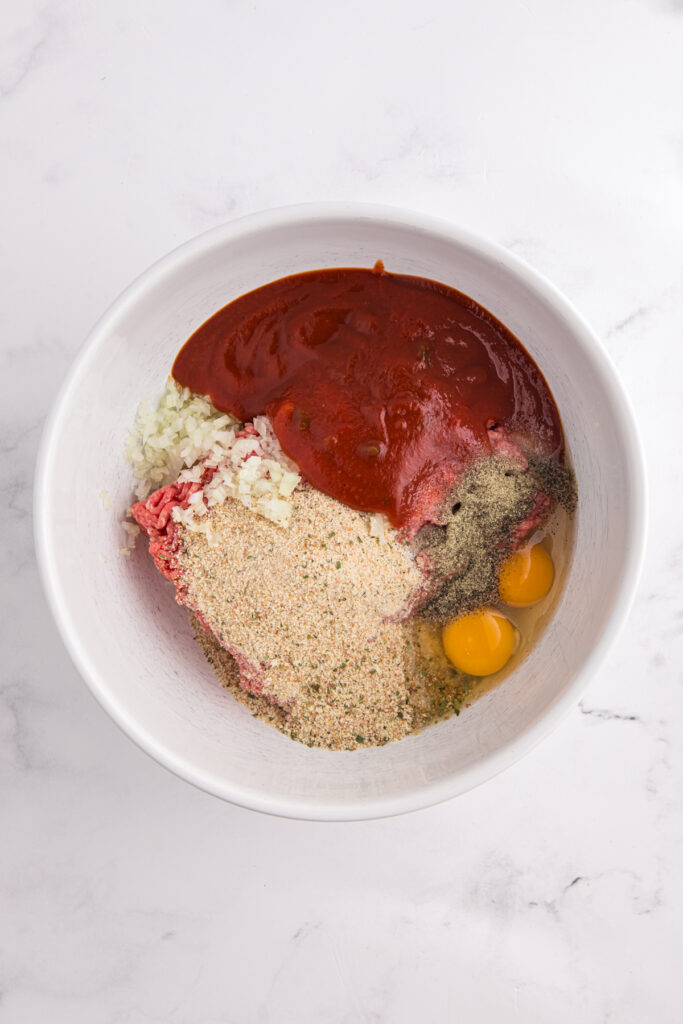 Top down image showing a white mixing bowl with a red sauce, eggs, breadcrumbs, seasoning, onion, and breadcrumbs