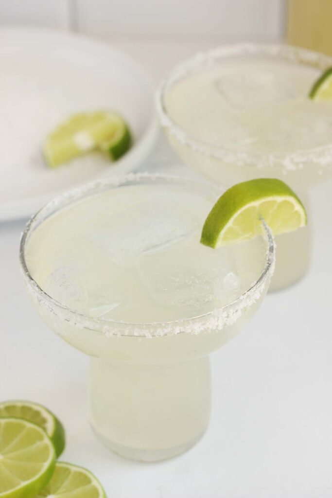 close up image of a margarita glass filled with ice and cocktail with a rimmed edge and lime wedges