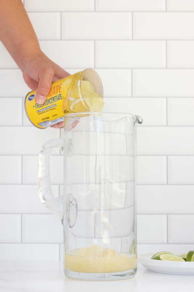 lemonade concentrate being poured into a pitcher with lime wedges off to the side