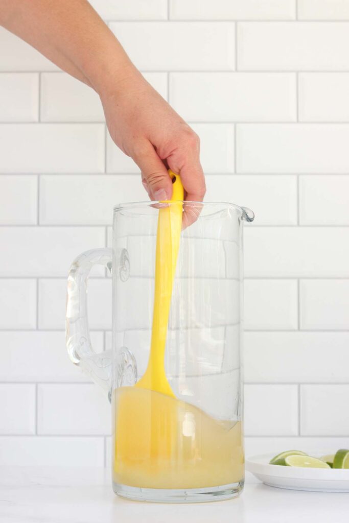 yellow spatula stirring liquid in a pitcher with lime wedges off to the side