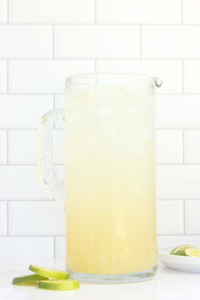close up image of a pitcher filled with margarita. Lime wedges are off to the side