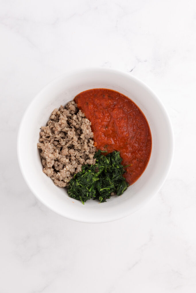 Top down image showing a white mixing bowl filled with sausage, pasta sauce, and spinach unmixed
