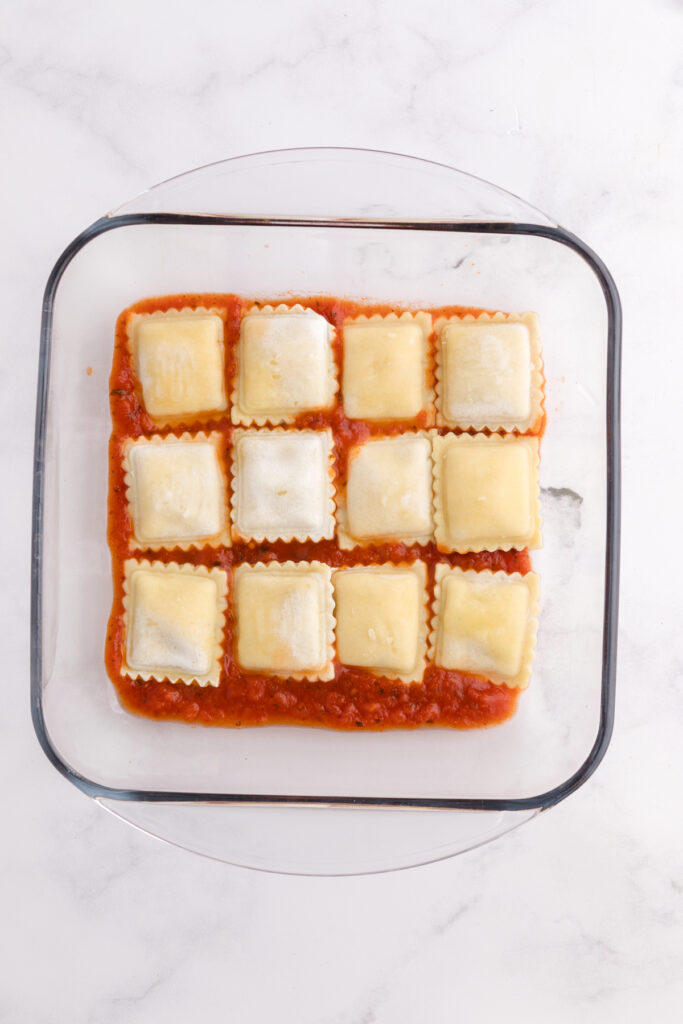 Top down image showing a glass square dish filled with pasta sauce topped with ravioli