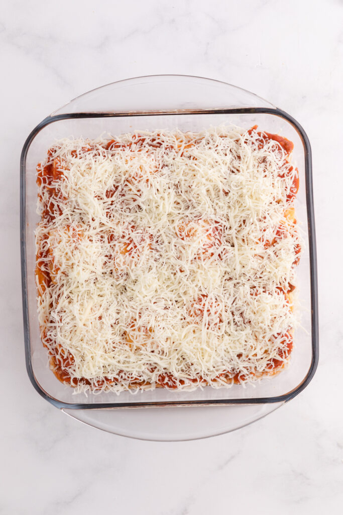Top down image showing a glass square dish filled with pasta sauce and beef topped with shredded cheese