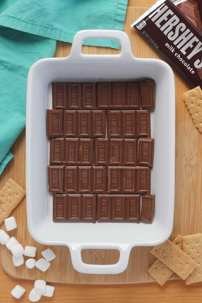Top down image showing hershey's chocolate bars broken into pieces layering the bottom of a white rectangle baking dish