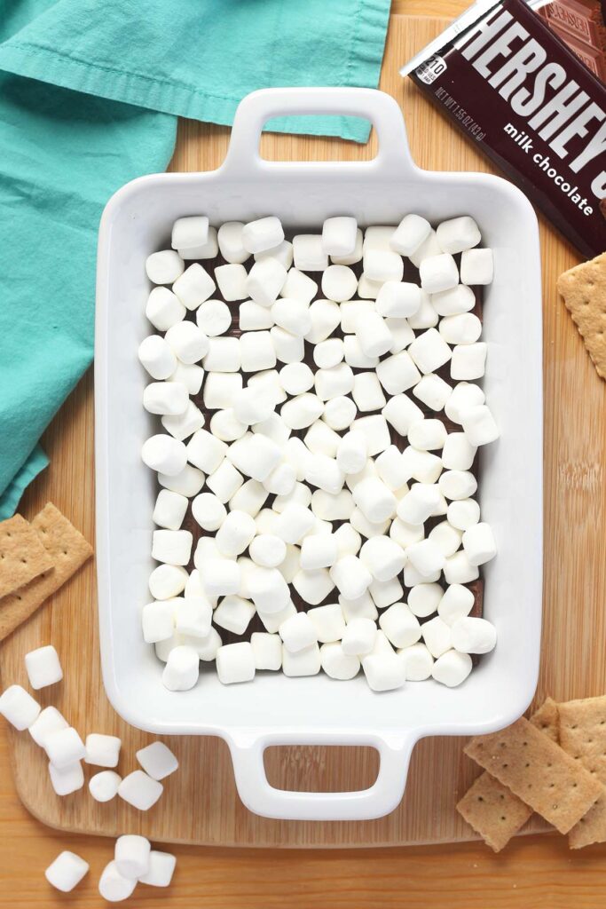 Top down image showing hershey's chocolate bars broken into pieces and mini marshmallows layering the bottom of a white rectangle baking dish