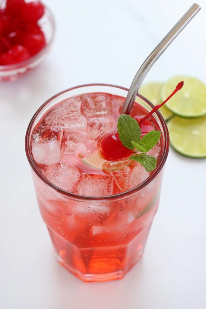 close up image showing a tall glass filled with a red drink that is topped with cherries, lime, and mint and a metal straw