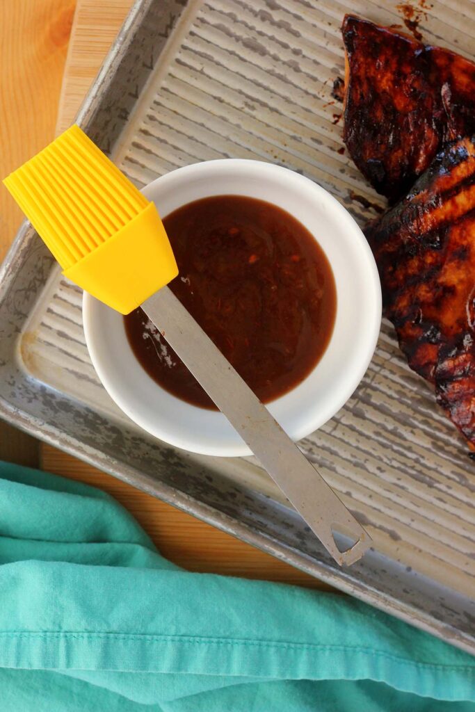 Top down image showing a white bowl on a serving tray filled with bbq sauce. A yellow silicone brush on top