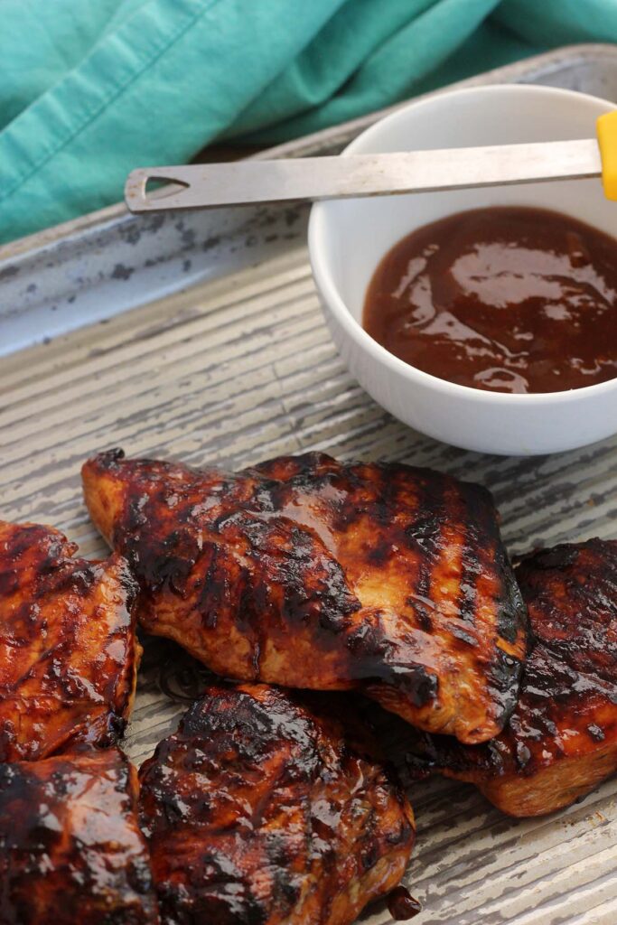 closeup image showing a tray of grilled chicken that has been brushed with a sauce. A bowl with more sauce and a silicone brush off to the side