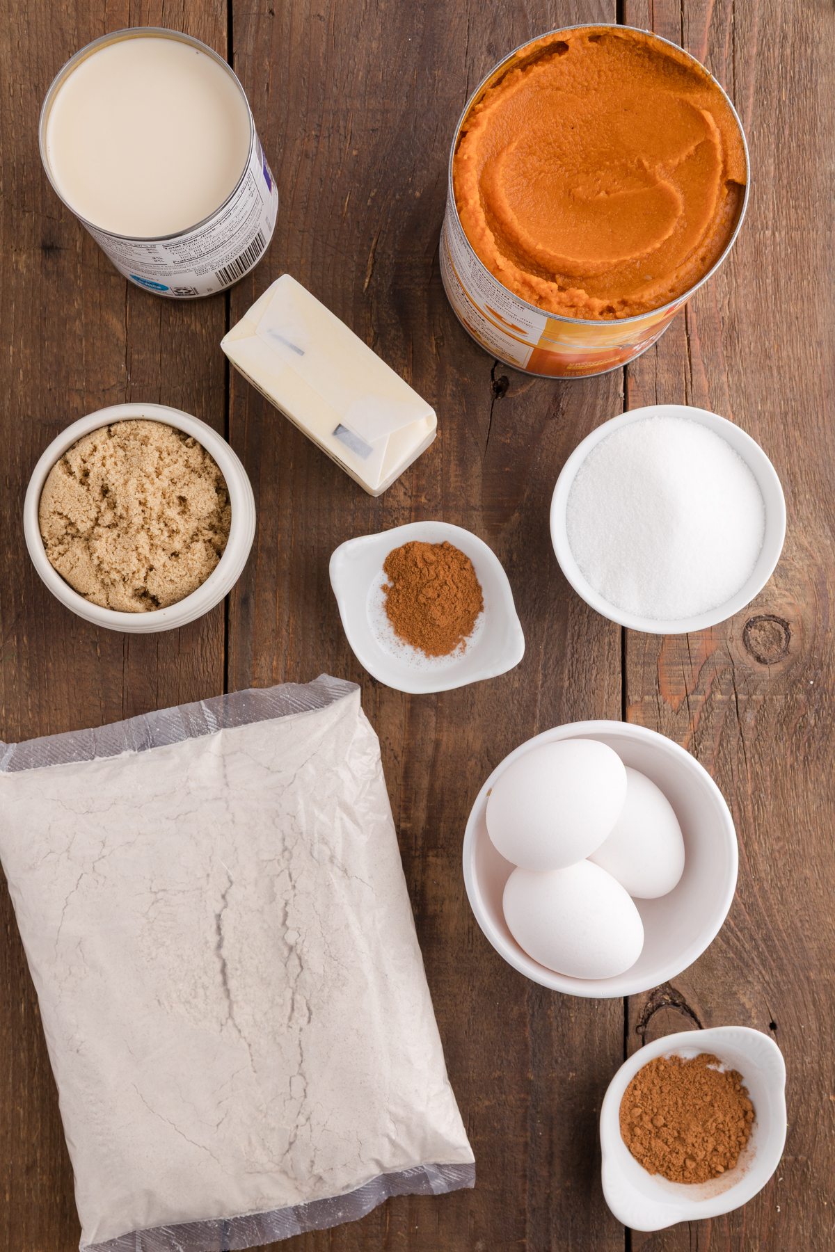 top down image showing a can of evaporated milk, a can of pumpkin puree, a stick of butter, a bowl of brown sugar, bowl of granulated sugar, dish with cinnamon, three eggs, and a cake mix