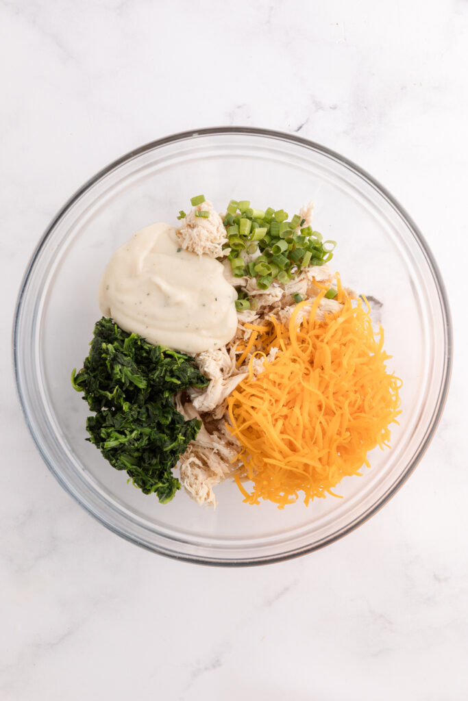 Top down image showing a glass mixing bowl filled with spinach, chicken, cheddar, green onion and a white sauce unmixed
