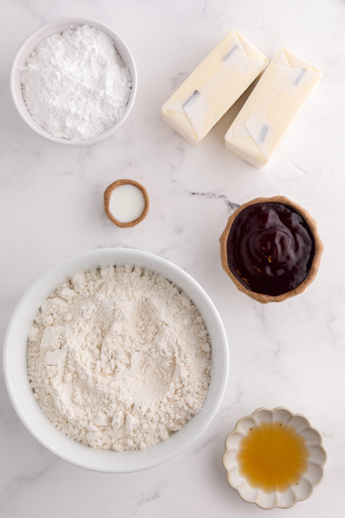 Top down image showing a bowl of flour, baking soda, powdered sugar, jam, butter and vanilla
