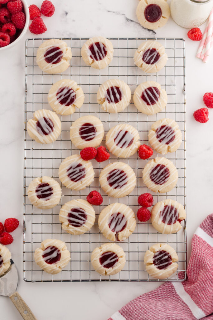Top down image showing a wire rack filled with cookies that have a raspberry jam filling and glaze drizzle. There are fresh raspberries around as well.