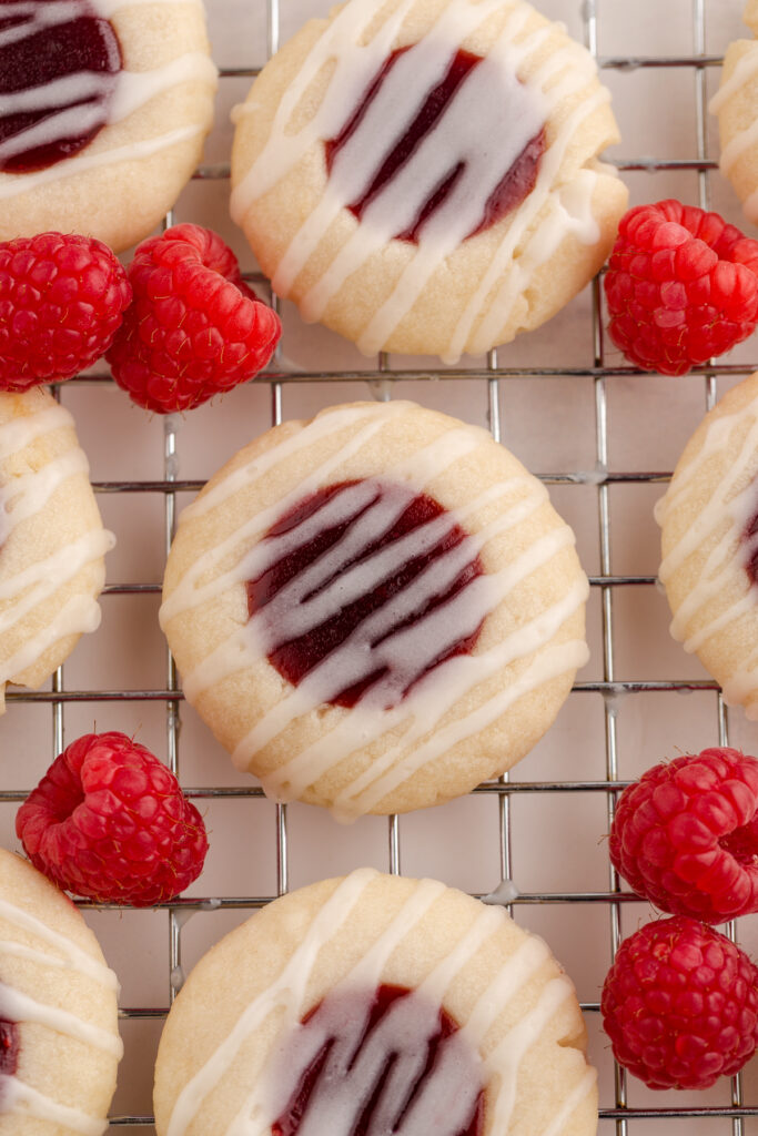 close up image showing small shortbread cookies filled with a raspberry jam and drizzled with a glaze. There are fresh raspberries around the cookies