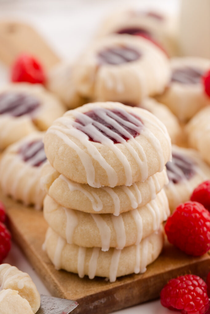 close up image showing a stack of four shortbread cookies with raspberry jam filling and glaze stacked on a tray with more cookies and raspberries