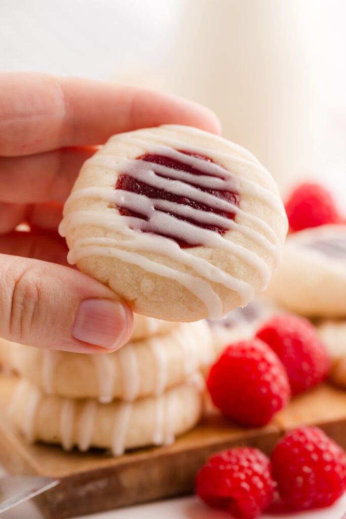 hang holding a single cookie that is filled with jam and a drizzle of glaze. Raspberries are off in the background with more cookies