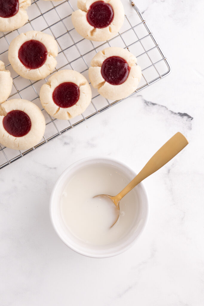 Top down image showing baked cookies with jam filling sitting on a wire cooling rack with a small bowl of glaze with a gold spoon inside