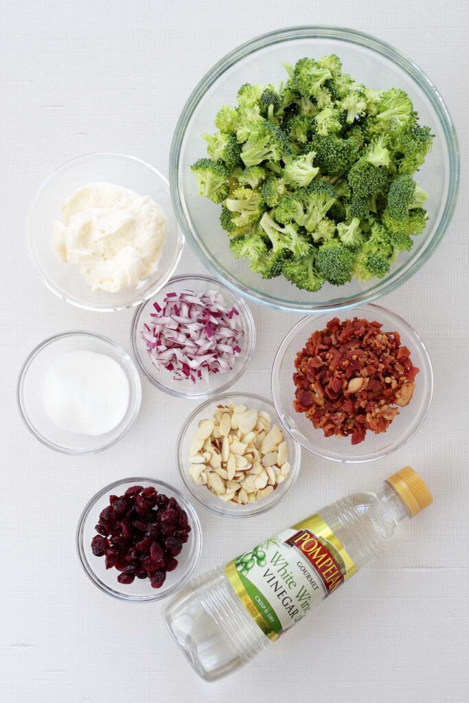 top down image showing a bowl of broccoli, a bowl of mayo, a bowl of red onion, bacon, sugar, almonds, craisins, and a container of white wine vinegar