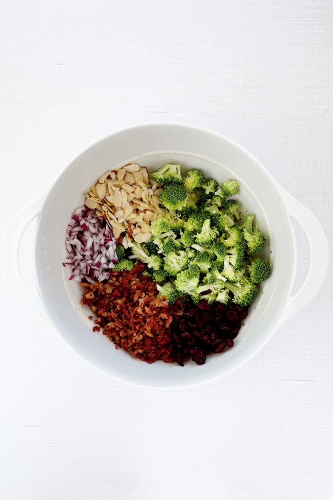 top down image showing a white mixing bowl with broccoli, almonds, red onion, bacon, and craisins unmixed