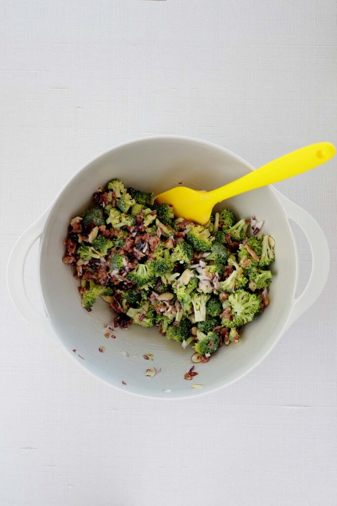 top down image showing a white mixing bowl with broccoli, almonds, red onion, bacon, and craisins mixed with a yellow spatula