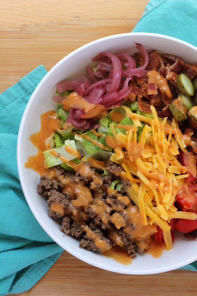 Top down image showing ground beef, lettuce, cheese, tomatoe, pickled onion and pickles in a white bowl with an orange sauce