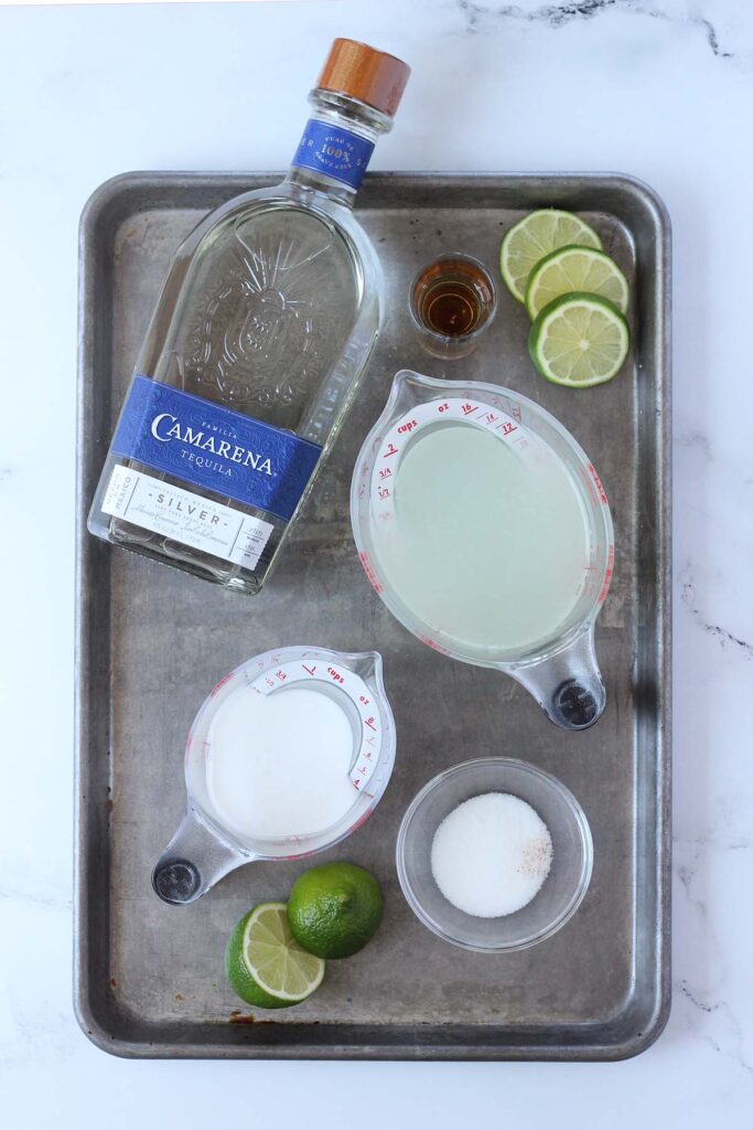 Top down image showing Camarena tequila, a glass of brown liquid, lime slices, two measuring cups with liquid, and a small glass bowl of sugar. 