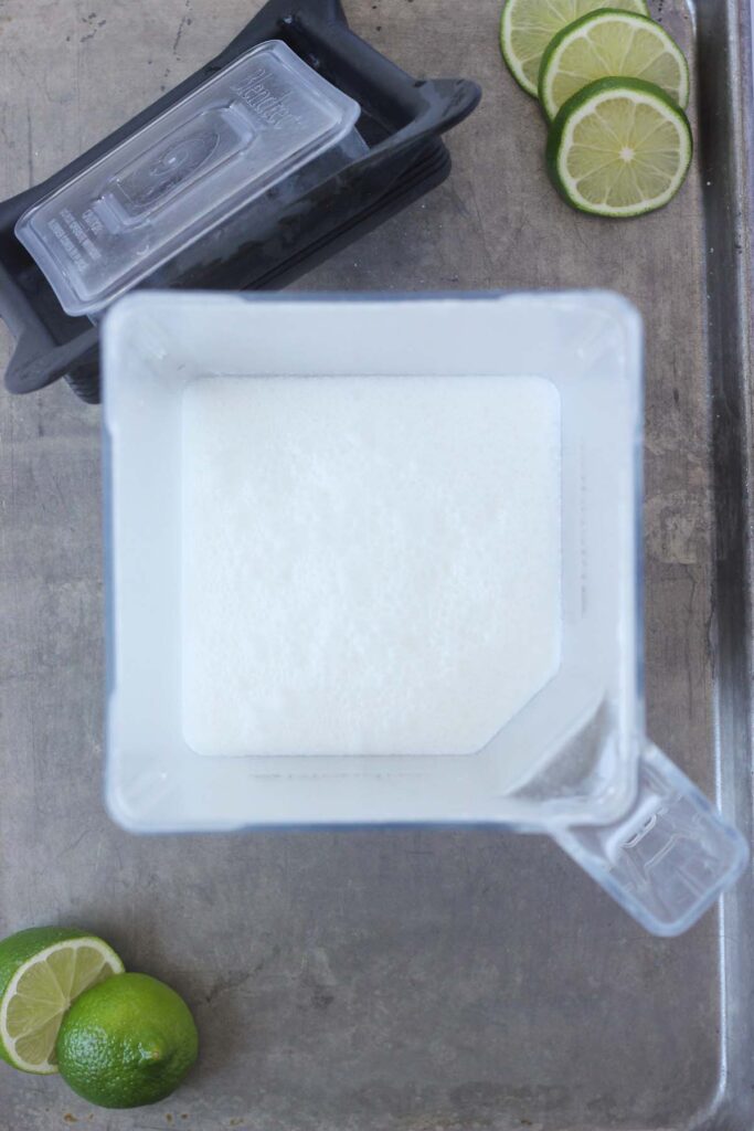 Top down image shownig white liquid inside of a blender. The lid of the blender and sliced limes off to the side