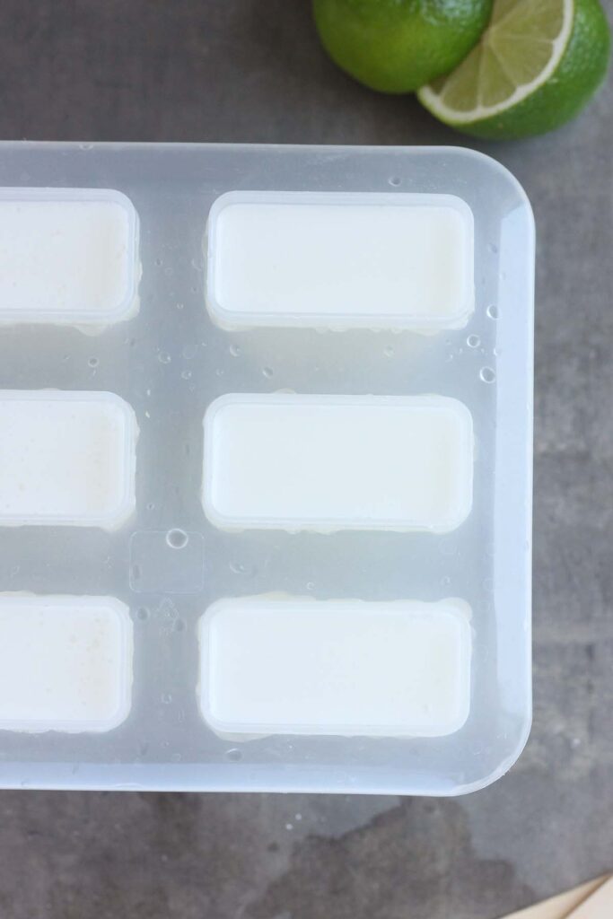 Top down image showing a close up of a popsicle mold filled with white liquid with lime slices off to the side