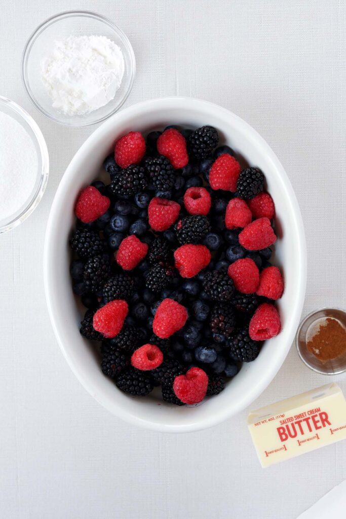 top down image showing an oval baking dish filled with blueberries, raspberries, and blackberries on a white table top with a stick of butter, small dish of cinnamon, small dish of sugar an done with cornstarch