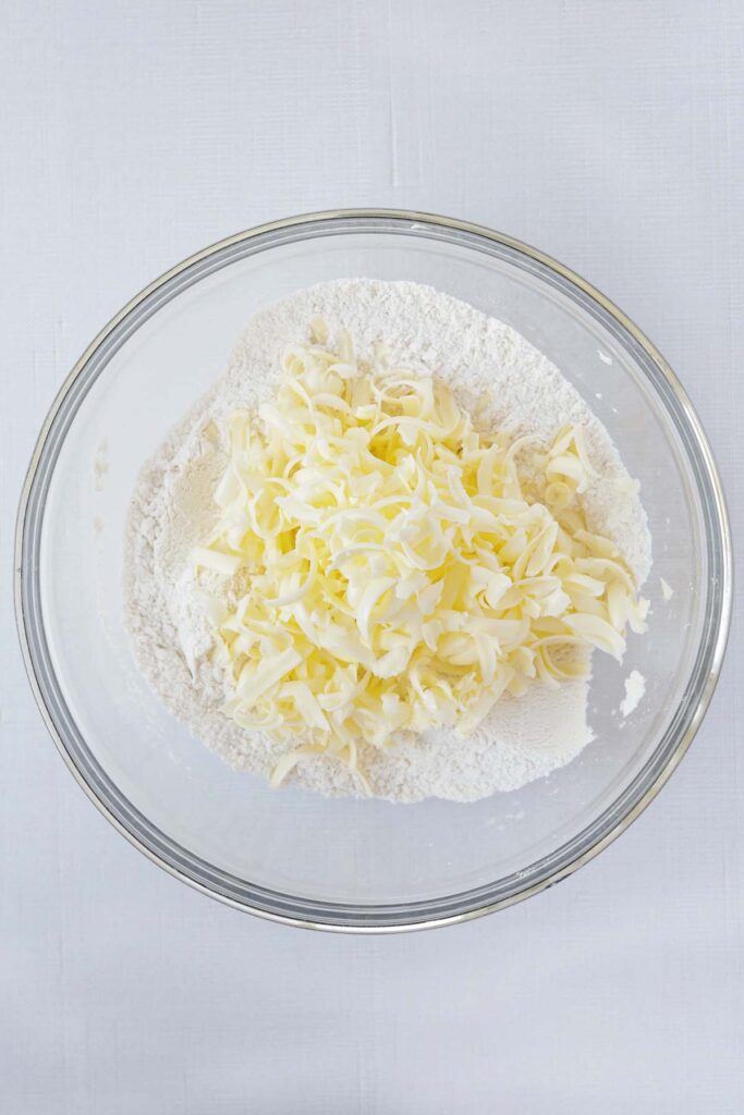 Top down image showing a glass mixing bowl filled with flour and shaved butter pieces on a white table