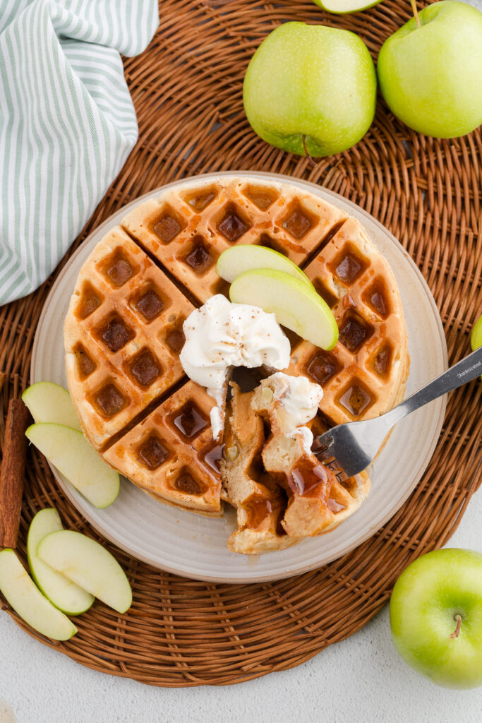Top down image showing an off white plate with a waffle. There is whipped topping and sliced apples on top with a bite sitting on a fork. Sliced apples are around the plate along with whole green apples