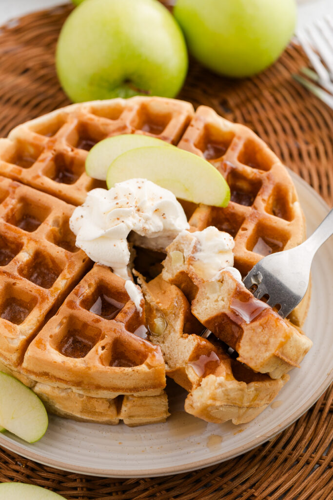 close up image showing a waffle covered in syrup, whipped topping and sliced apples with a bite on a fork.