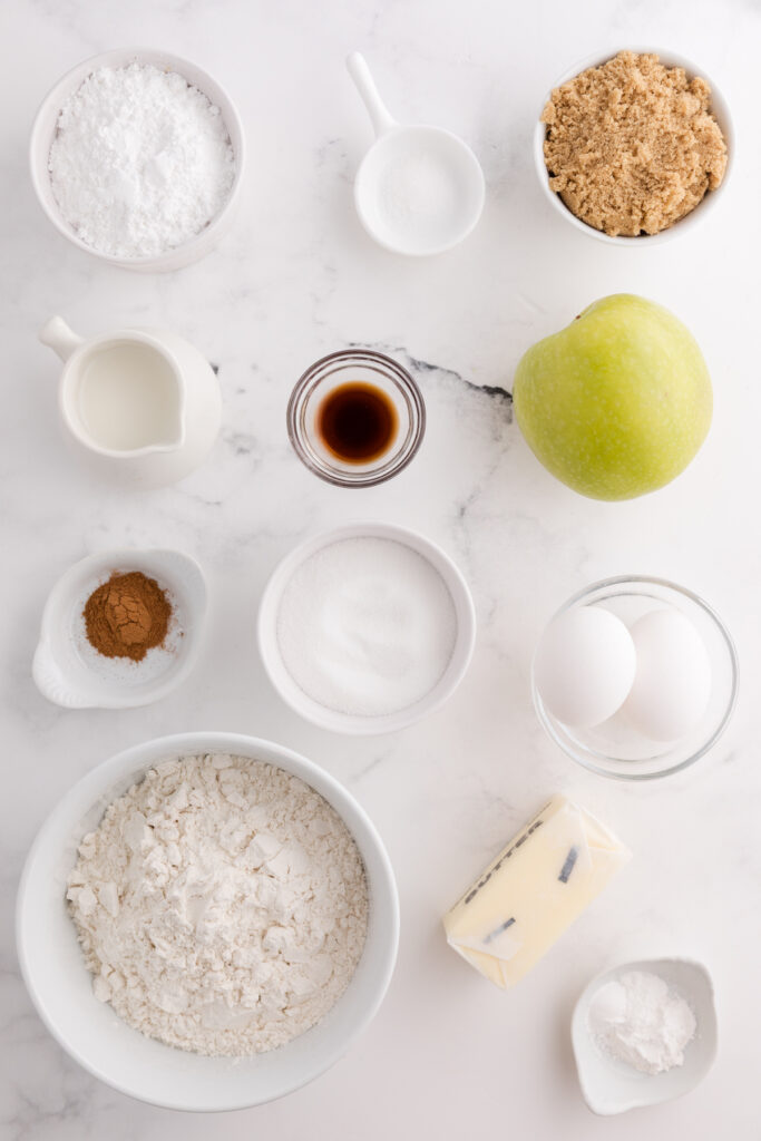 Top down image showing all of the ingredients you would need to make an apple bread divided out into their own bowls on a white table top