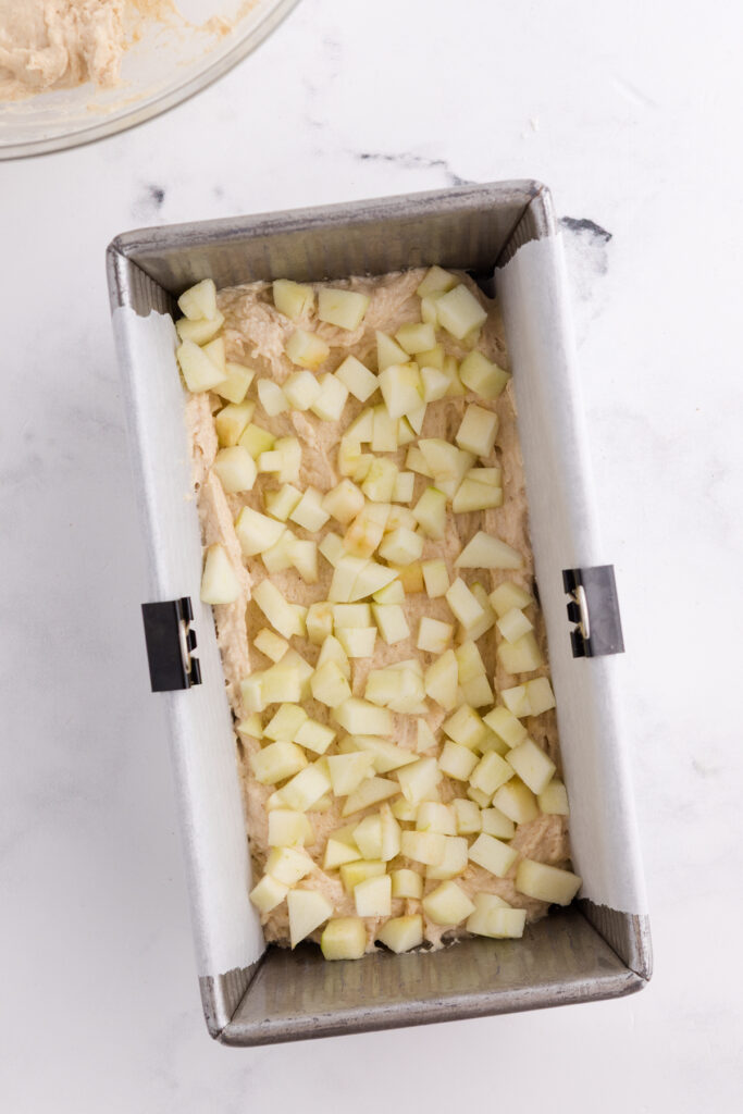 Top down image showing a loaf pan that has a parchment paper and batter inside topped with chopped apples