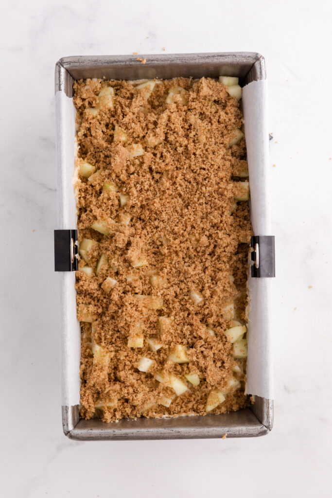 Top down image showing a loaf pan that has a parchment paper and batter inside topped with chopped apples and a cinnamon sugar mixture