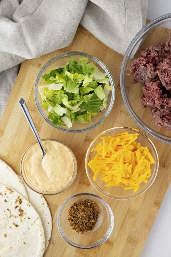 Overhead view of wrap ingredients on a wooden board, including shredded lettuce, raw ground beef, shredded cheddar cheese, tortillas, creamy special sauce in a small bowl with a spoon, and a small bowl of seasoning blend.