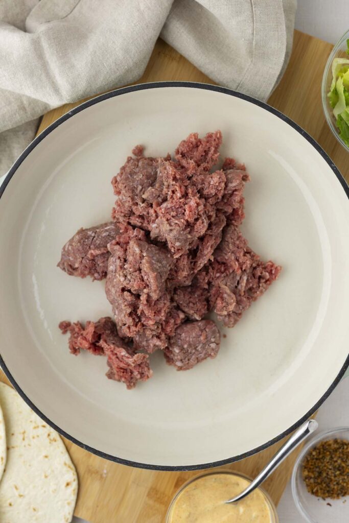 Raw ground beef in a large white skillet, surrounded by ingredients for Mac wraps, including tortillas, special sauce, seasoning, and shredded lettuce on a wooden board.