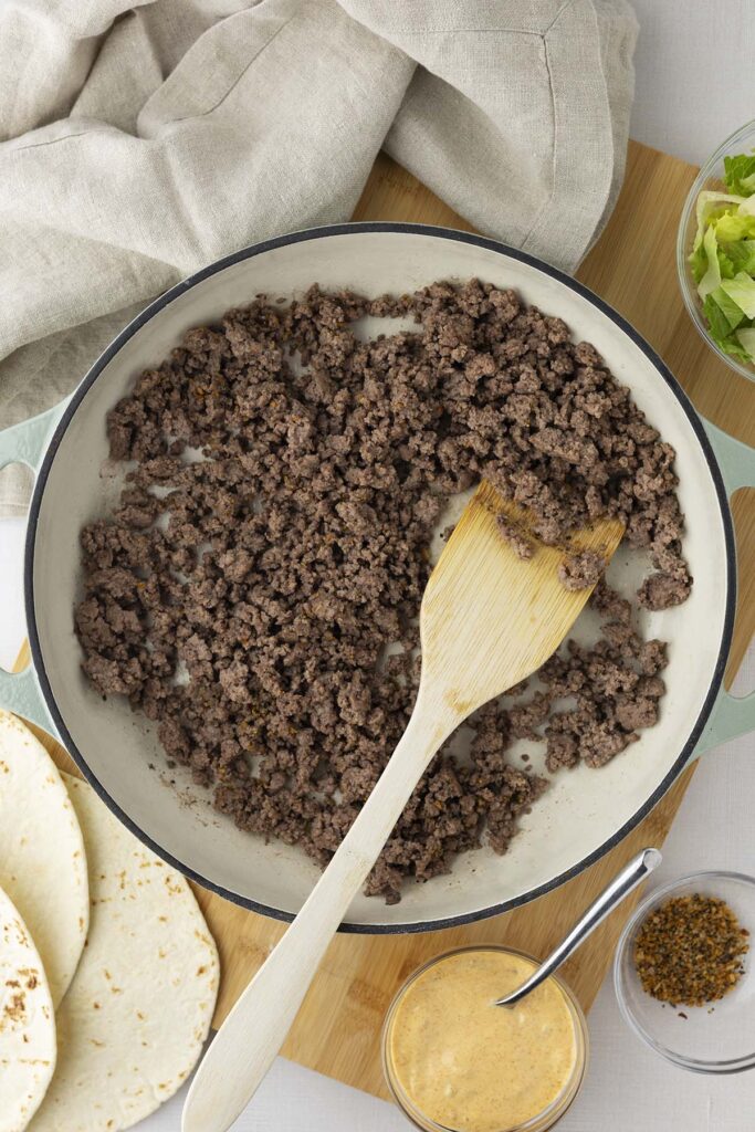 Ground beef in a large white skillet, surrounded by ingredients for Mac wraps, including tortillas, special sauce, seasoning, and shredded lettuce on a wooden board.