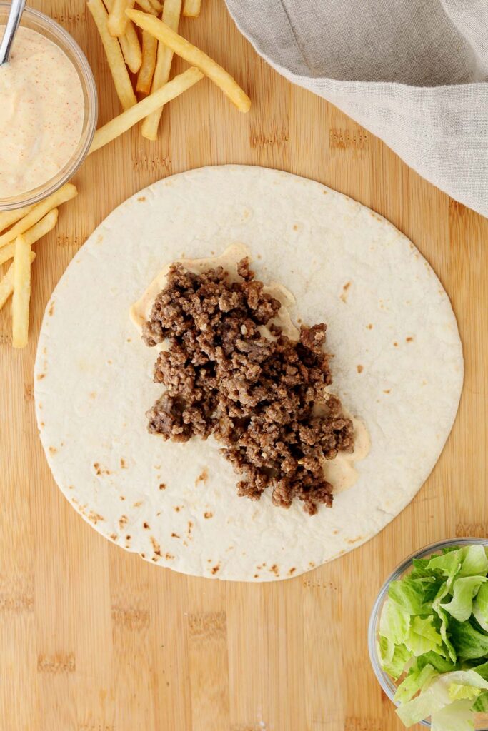 Top down image showing a tortilla on a wooden table top with a burger sauce spread on top with ground beef