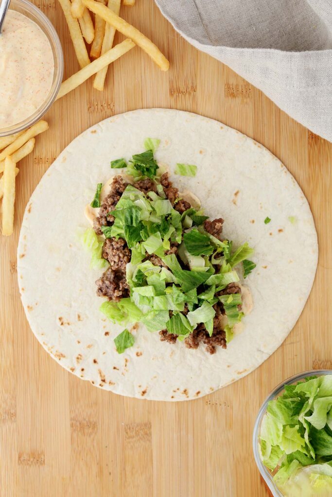 Top down image showing a tortilla on a wooden table top with a burger sauce spread on top with ground beef and lettuce