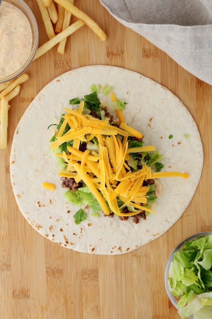 Top down image showing a tortilla on a wooden table top with a burger sauce spread on top with ground beef and lettuce sprinkled with cheese