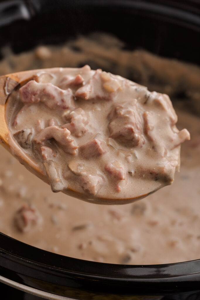 Close up image showing a spoon scooping out a creamy beef mixture from the inside of a slow cooker