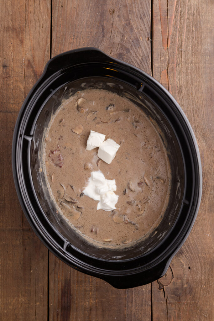 Top down image showing a creamy beef mixture inside of a slow cooker with dots of cream cheese block on top