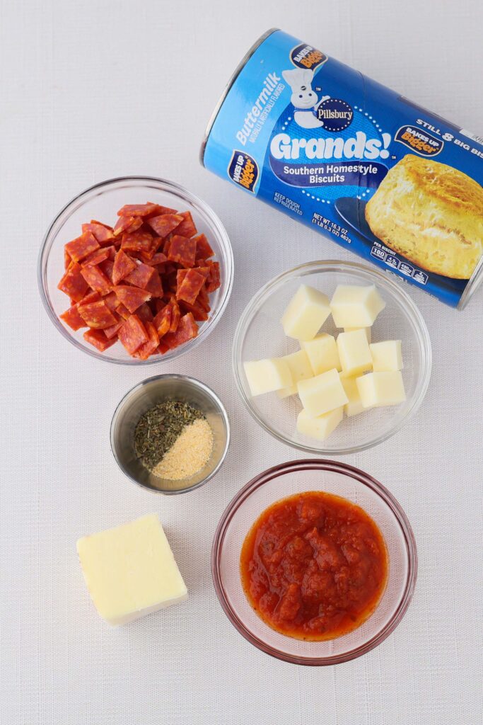 top down image showing a can of biscuits with a bowl of pepperoni pieces, a bowl of mozzarella cubes, a dish with seasonings, a pat of butter and a bowl of pizza dough